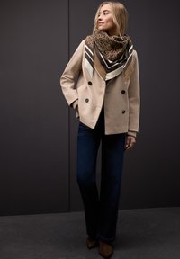 Veste beige croisée à boutons noirs, associée à un jean évasé bleu marine et à une écharpe marron à motifs avec des rayures et des touches d'imprimé léopard.