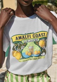 Valge T-särk, millel on värviline Amalfi rannikul asuvate sidrunite graafika, kollased sidrunid, rohelised lehed ja klaasi illustration. Näha on triibulised rohelised püksid.