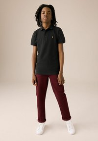 Polo noir à manches courtes, conçu avec un petit logo jaune ; associé à un pantalon bordeaux et des baskets blanches.