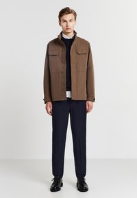 Giacca leggera marrone con due tasche sul petto, chiusura a bottoni e colletto rialzato, abbinata a pantaloni navy a trama e scarpe nere.