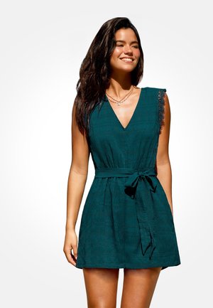 Donna con capelli lunghi e scuri che indossa un abito verde scuro senza maniche con bordi in pizzo e vita annodata, sorridente su sfondo bianco.