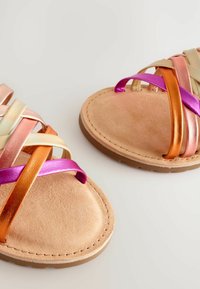 Zwei flache Sandalen mit breiten Riemen in metallischen Farbtönen von Pink, Orange und Gold, auf einem beigen Wildlederfußbett mit einer strukturierten Sohle.