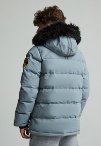 Veste rembourrée bleu clair avec une capuche en fausse fourrure noire, un design matelassé et un patch sur la manche gauche. Rayures grises à l'arrière.