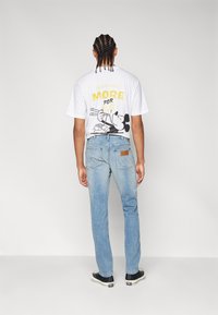 Bela grafična majica s kratkimi rokavi z dizajnom Mickey Mouse in rumenim napisom na hrbtni strani, v kombinaciji s svetlo modrimi ravnimi jeans hlačami in črnimi športniki.