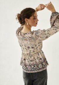 Top de peplum con estampado floral en rosa claro, con mangas largas y abullonadas, que presenta una cintura ceñida y un dobladillo más oscuro en contraste. Llevado con jeans oscuros.