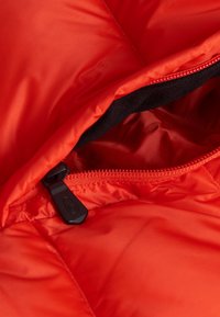 Rode geïsoleerde jas met een gestructureerd oppervlak, uitgerust met een zwarte rits en een gladde voering. Zichtbaar ritsdetail bij de opening.