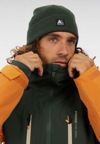 Dunkelgrüne Jacke mit orangefarbenen Akzenten und Reißverschlüssen. Mit Fleece gefütterter Kragen, strukturiertes Material und Logo-Patch. Gestrickte grüne Mütze.