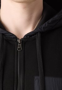 Detailní záběr na černou mikinu s kapucí na zip s tkaničkami, která je oblečena přes béžové tričko, zobrazuje krk a bradu osoby.