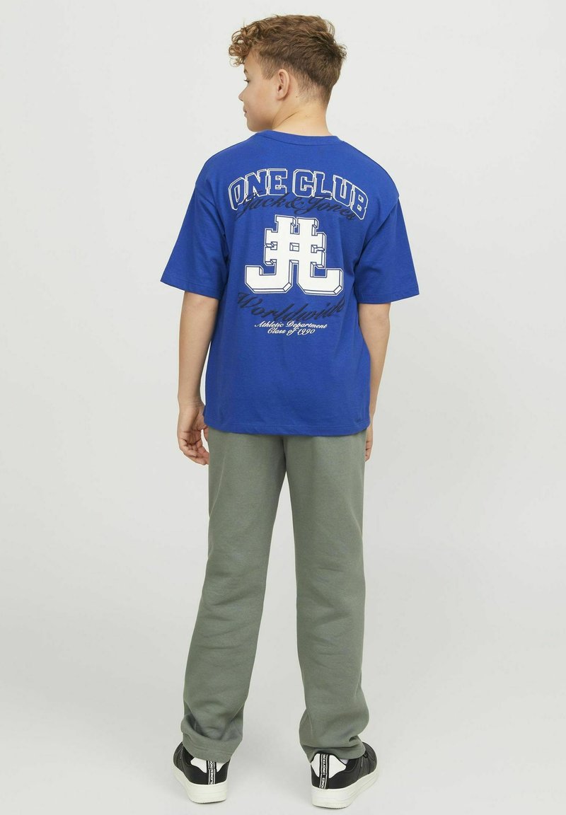 Blauwe katoenen t-shirt met een grote witte graphic op de achterkant, met tekst en een logo. Gecombineerd met grijze joggingbroek en zwarte sneakers.