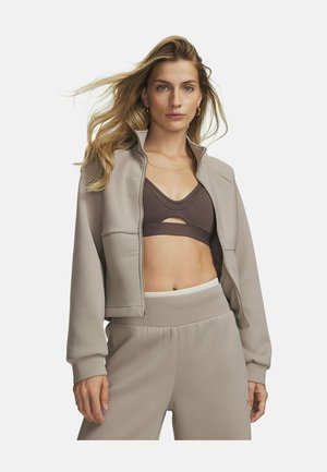 Vrouw met lang blond haar die een beige jas met rits open draagt over een donkere sportbeha en bijpassende beige hoogtaillebroek, staand tegen een witte achtergrond.