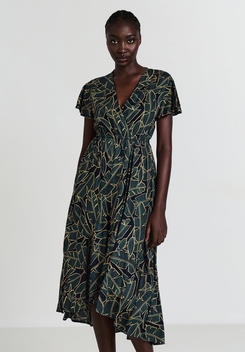 Robe portefeuille en tissu avec une base vert foncé et un motif de feuilles dorées. Présente des manches flottantes et une taille cintrée pour une silhouette fluide.