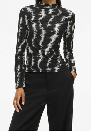 Kobieta ubrana w czarną bluzkę z długim rękawem z białym abstrakcyjnym wzorem zygzaków, w połączeniu z luźnymi czarnymi spodniami z wysokim stanem, ręka w kieszeni.