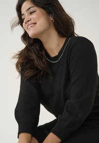 Black knit sweater with a round neckline, ribbed cuffs, and a smooth texture. Worn with a delicate silver chain necklace.