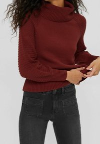 Pull en maille bordeaux avec un col roulé. Doté de manches texturées et de poignets côtelés. Associé à un jean foncé.