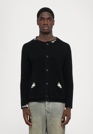 Mand iført en sort knaplukket cardigan med hvide mønsterdetaljer på kraven, lommerne og mansjetterne, parret med lysegrå jeans.