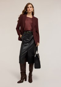 Donna vestita con un blazer bordeaux e top, gonna midi in pelle nera, stivali al ginocchio con stampa pitone, mentre tiene una borsa nera.