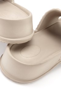 Beige Clogs aus einem strukturierten, gummiähnlichen Material. Sie verfügen über eine abgerundete Zehenpartie, erhöhte Ränder und kreisförmige Akzente im Fußbett. Geeignet für die Freizeit.