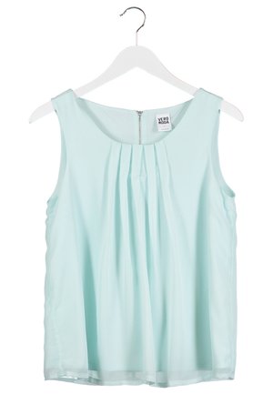 Kobieta ubrana w białą bluzkę bez rękawów, ciemnoniebieskie podwinięte dopasowane jeansy oraz czarne czółenka w szpic, niosąca czarną torebkę na łańcuszku.