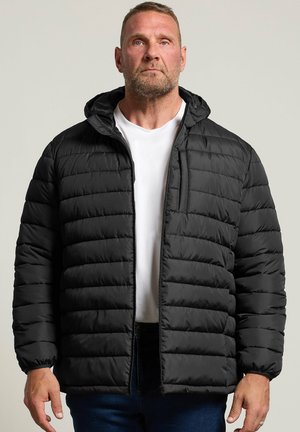 Homme portant une veste matelassée noire par-dessus une chemise blanche et un jean bleu, debout devant un fond clair uni.