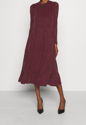 Frau trägt ein bordeauxfarbenes Midikleid mit langen Ärmeln, Rüschensaum und schwarzen spitzen Schuhen, steht vor einem schlichten Hintergrund.