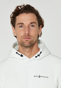 Sudadera blanca con capucha ajustable, logo impreso "SAIL RACING" en el pecho, con detalles en negro y tela texturizada.