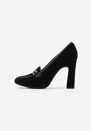 Zapato de tacón alto de ante negro con punta redondeada, acentos de cuero decorativos y un detalle en hardware dorado en la parte delantera.