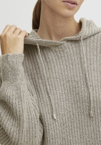 Beige Kapuzenpullover mit gerippter Textur, lockerer Passform und Kordelzügen mit silbernen Spitzen. Das weiche Material betont den Komfort.