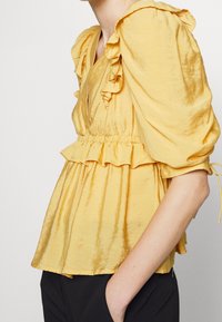 Blusa in tessuto giallo sgualcito con maniche arricciate e una vita con elastico. Il design include un dettaglio a peplum.