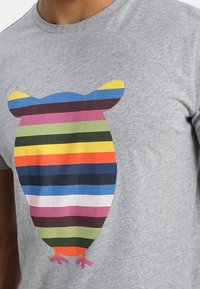 Hombre que lleva una camisa gris con un gráfico de un búho a rayas multicolores centrado en el pecho.