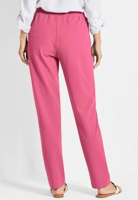 Roze stoffen broek met een zachte textuur, taps toelopende pijpen en een elastische tailleband. Draagt bij sandalen, gezien vanuit de achterkant.