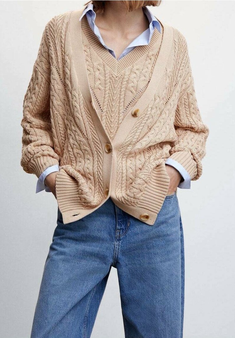 Cardigan beige en tricot avec motifs torsad és texturés, coupe oversize et poignets côtelés. Comprend deux gros boutons et une chemise bleue superposée en dessous.