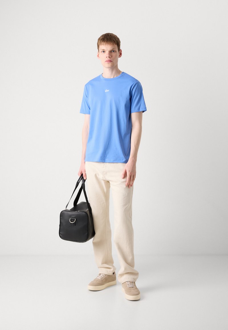 Niebieska koszulka z okrągłym dekoltem z napisem "Good", beżowe spodnie o szerokich nogawkach, beżowe trampki oraz czarna torba na zamek z metalowym akcentem.