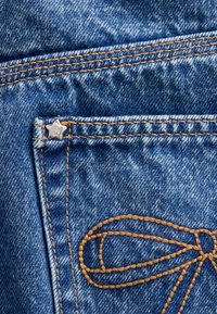 Denimstof met een blauwe tint, voorzien van gele stiksels. Inclusief een stervormig metalen label en een gedetailleerd contrasterend geborduurd ontwerp.