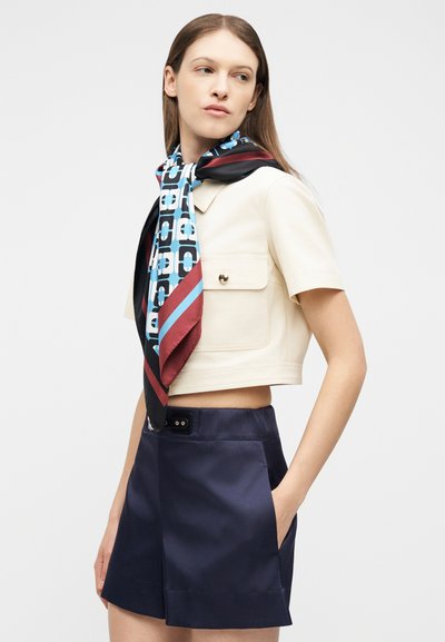 Γυναίκα που φοράει εκρού κοντό σακάκι με κοντά μανίκια, ναυτικό σορτς με ψηλή μέση και μεταξωτό φουλάρι μπλε, μαύρου και μπουργκούντι με σχέδιο γύρω από το λαιμό της.