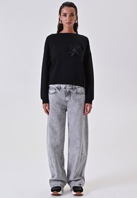 Felpa nera con un accento floreale sul petto, abbinata a jeans a gamba larga grigio chiaro con una texture sbiadita. Sneakers bianco e nere.