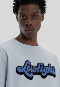 Ανοιχτό μπλε βαμβακερό t-shirt με μια υφή, μαύρη και μπλε κεντημένη γραφική παράσταση "Lowlights" στο μπροστινό μέρος. Κοντά μανίκια, στρογγυλή λαιμόκοψη.