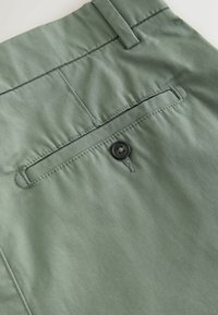 Gros plan de la poche arrière d’un pantalon vert olive avec fermeture à bouton et passant de ceinture sur tissu lisse.