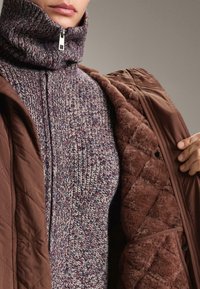 Pleteni pulover s visokim ovratnikom v burgundski in kremni barvi, oblečen pod rjavim prešitim jopičem s mehkimi, teksturiranimi podlogami.