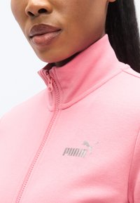 Ροζ ζακέτα PUMA με φερμουάρ, ψηλό γιακά, απαλό ύφασμα και ασημένιες λεπτομέρειες λογότυπου στην αριστερή πλευρά. Κοντινή όψη του λαιμού και της λεπτομέρειας.