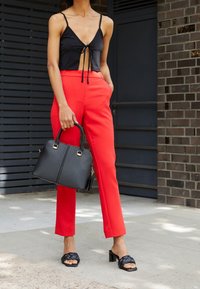Žena v černém tílku se zavazováním vpředu, jasně červených kalhotách s vysokým pasem, černých sandálech na podpatku, držící černou kabelku.