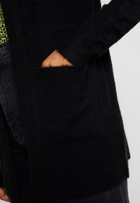 Svart stickad cardigan med långa ärmar, med en framficka och ribbade manschetter. Texturen ser mjuk och lurvig ut.