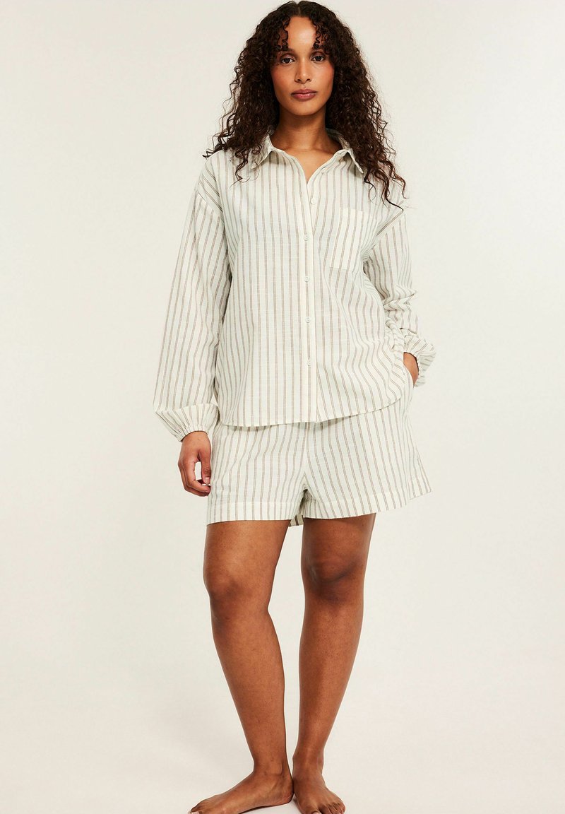 Kvinde med krøllet hår iført beige og hvide lodrette stribede skjorte med lange ærmer og matchende shorts, stående barfodet mod en ensfarvet baggrund.