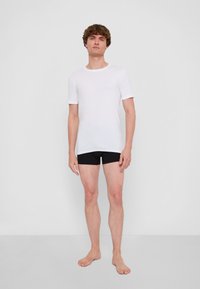 T-shirt bianca a maniche corte dal design aderente, indossata con boxer neri. Texture liscia, stile minimalista contro uno sfondo semplice.
