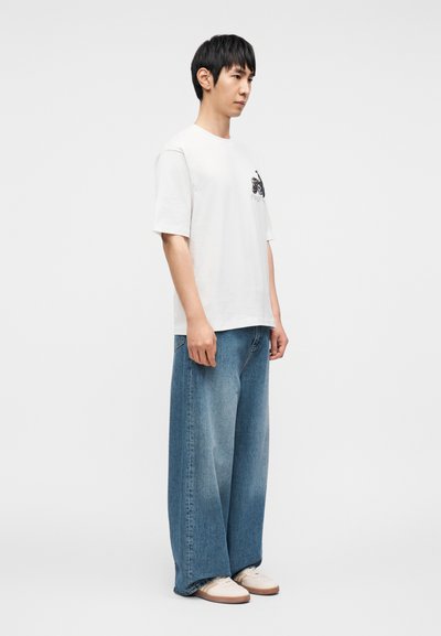 Λευκό κοντομάνικο μπλουζάκι με μαύρο γραφικό στο στήθος, συνδυασμένο με φαρδιά, ανοιχτό μπλε τζιν παντελόνια με ξεθωριασμένη υφή.