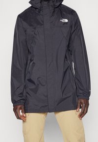 Svart vattentät jacka med huva, som har en framzipper, elastiska ärmslut och The North Face-logotyp på bröstet. Slätt, lätt material.