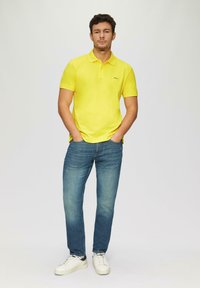 Gelbes Poloshirt mit klassischem Kragen, kombiniert mit blauen Jeans und weißen Sneakers. Einfaches Design, glatte Textur, lässige Passform.