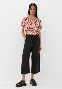 Blumenbluse in Rosa und Schwarz mit kurzen, schwingenden Ärmeln und einem Schlüsselloch-Ausschnitt, kombiniert mit schwarzen, weiten Hosen und Plateau-Sandalen.