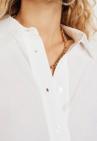 White button-up shirt featuring a classic collar, subtle texture, and small cut-out detail near the neck. Accented with a gold chain necklace.