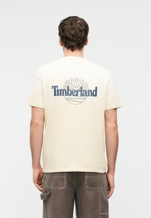 Homme portant un T-shirt Timberland beige avec un logo bleu et un motif d'arbre dans le dos, associé à un pantalon cargo marron, tourné de dos.