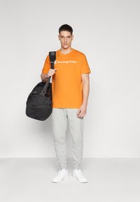 Oranges Baumwoll-T-Shirt mit "Champion"-Logo, graue Jogginghose, schwarze Sporttasche und weiße Sneakers. Lässiges Sportoutfit.